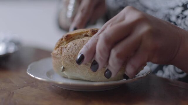 A woman's hand with painted nails cuts bread with a knife starting from the very edge. The bread is called Marraqueta, a traditional bread from La Paz, Bolivia.