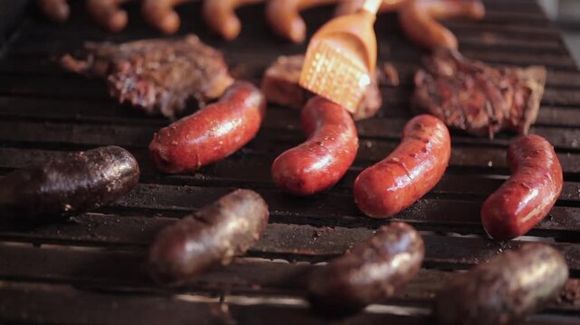 Seasoning the chorizo on the grill. On a grill, there is a variety of meats and blood sausage, while a brown plastic spatula seasons the orange sausage.