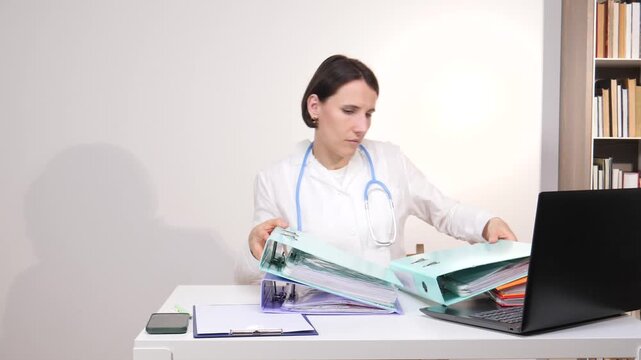 Medical professional navigating through multiple patient files and folders, organizing paperwork at a desk with a laptop, highlighting the administrative workload in healthcare