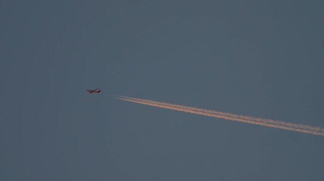 A commercial aircraft flies at high altitude trailing two parallel contrails lit golden by the setting sun against a cool blue-grey evening sky