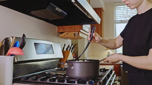 A person stands in a kitchen stirring food in a pot on the stove. Sunlight comes through the window, creating a bright atmosphere. The kitchen is organized and functional.