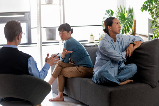 Two Asian women attending counseling session during emotional relationship conflict. One partner appears upset while therapist guides conversation about communication and trust