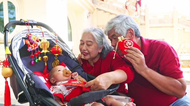 Grandparents took their infant grandson celebrate Chinese New Year visiting Chinese temple and playing with him in a stroller using Chinese doll decorate the stroller and shaking red envelopes as toy.