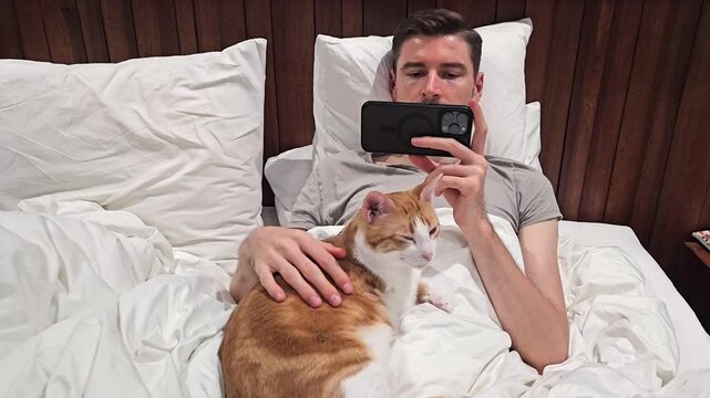 Young man lying in bed with orange and white cat while using smartphone. Cozy indoor lifestyle scene with pet companionship, mobile entertainment and relaxing evening at home. Front view close up