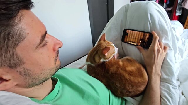 Man lying on bed with orange cat while watching video on smartphone. Relaxed home lifestyle scene with pet companionship, mobile entertainment and indoor leisure activity. Over shoulder view indoors