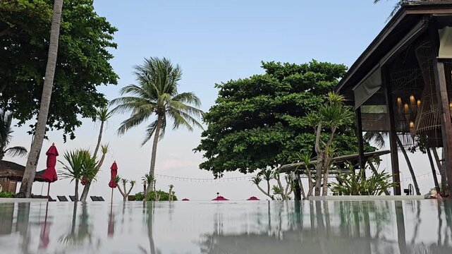 Tropical resort swimming pool with palm trees and lounge area at sunset, calm vacation landscape with reflections on water and coastal hotel architecture