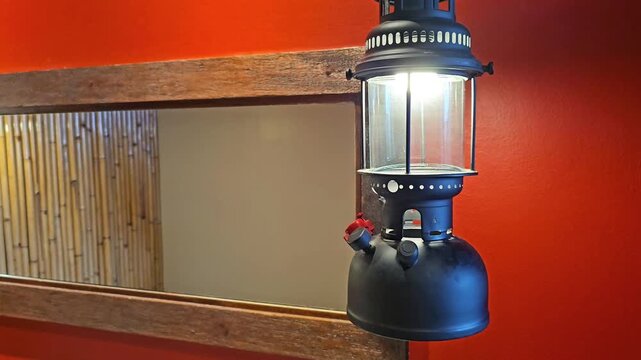 Black camping lantern mounted beside wooden framed mirror on orange wall interior. Illuminated vintage lamp with reflection and bamboo texture in rustic home decor setting. Static close up shot