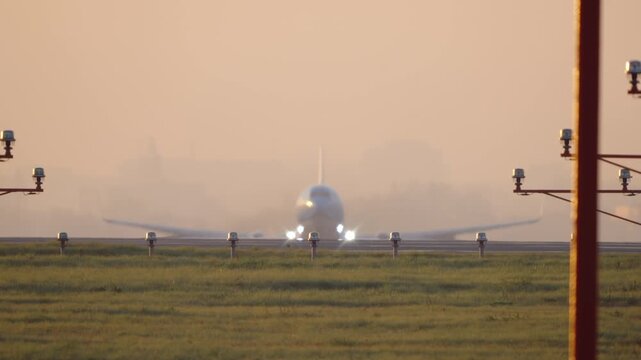 Passenger airplane emerging from glowing sunrise haze during takeoff, head on telephoto view with soft golden morning light and runway lights, calm cinematic aviation moment