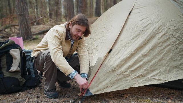 Male backpacker setting up camping tent in forest. Caucasian traveler preparing temporary shelter during outdoor expedition. Hiker organizing campsite and enjoying active tourism lifestyle in woodland