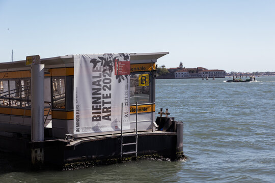 Venice, Italy 04 30 2026: A large Biennale Arte exhibition banner hangs on a yellow vaporetto water bus stop in the Venetian lagoon. A boat travels on the water under a clear blue sky. 
