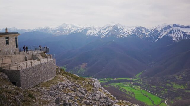 Breathtaking panorama of a high altitude mountain landscape where tourists are hiking on a path surrounded by patches of snow, with a stunning view of a distant, snow covered mountain range