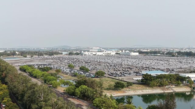 Aerial reveal shot through treetops unveiling vast parking lot filled with rows of new cars at automobile manufacturing facility