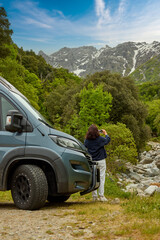 Active woman traveler standing beside a camper van and taking a photo of a mountain valley with forest and rocky peaks in Corsica. Colors: green, gray, blue. © Martin Mecnarowski