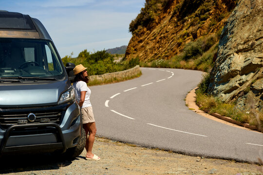 Middle aged woman traveler standing beside a camper van on a winding coastal mountain road during a road trip in Corsica. Colors: gray, blue, beige.