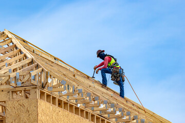 Low-angle view of a construction worker wearing a safety harness and hard hat using a hammer to install wooden roof trusses at a residential building site, USA © Andrei Liashchou