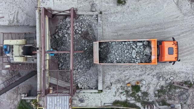 A truck unloads gravel at a construction site. Heavy machinery operates nearby, moving materials. Dust is present as workers are engaged in ongoing construction activities.