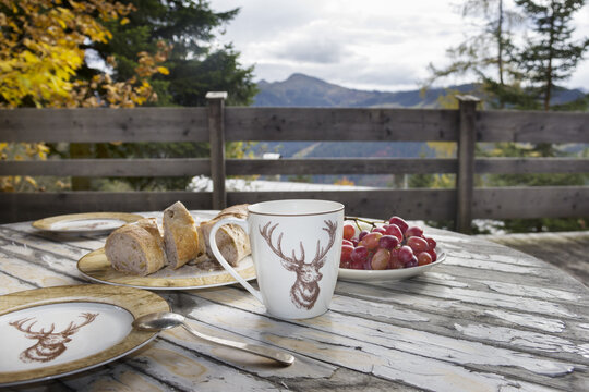 Cozy alpine breakfast on a wooden terrace with a mountain view. Mug with a deer print, fresh pastry, and red grapes on a rustic table.