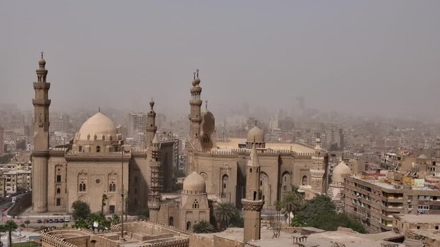 Two grand Cairo mosques with multiple minarets and imposing domes rise above the sprawling hazy cityscape of Egypt's capital, a symbol of Islamic heritage and history.