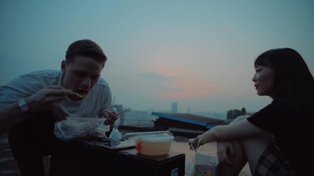 Young man and woman eating takeout food and talking on a city rooftop during a beautiful sunset