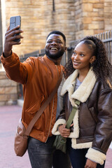 African American couple taking smartphone selfie, laughing on street in coats, crossbody bags © wavebreak3