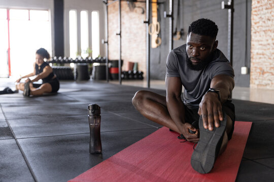 Diverse gymmates stretching on rubber-tiled floor at gym with red mat, water bottle, smartwatch