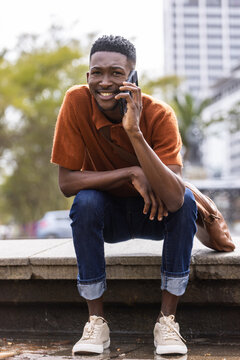 Man crouching on stone ledge by water at plaza, holding phone, rust polo, backpack beside