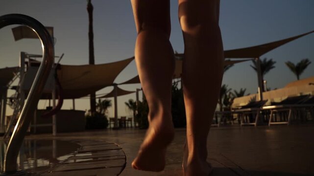 Sunset glows behind poolside lounge chairs. Feet walk barefoot, steps echo softly on deck. Silhouettes frame a calm, warm evening atmosphere. No emotion visible, only quiet movement