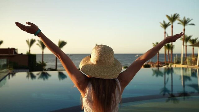 Woman relaxes by infinity pool, arms raised. Sunset glows over ocean and palm trees. She wears straw hat, feels calm and free. Perfect vacation moment, serene and joyful. Ideal for travel, wellness