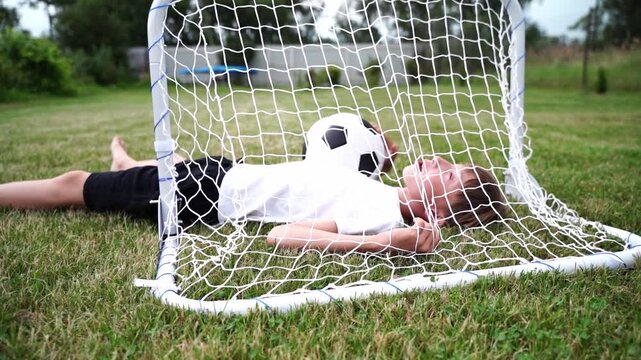 Child rests inside soccer goal net, smiling. Ball nestled close, playful moment captured. Green grass, open sky, gentle outdoor setting. Perfect for kids' sports or playful leisure scenes