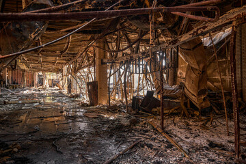 Fototapeta premium A long view of an industrial corridor filled with hanging wires and collapsed metal ventilation ducts after a fire. Water and debris cover the ground of the ruined facility.