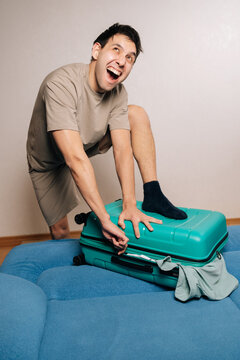 Vertical portrait of angry traveler male struggling to zip overpacked green suitcase for a trip, frowning and making a funny face, experiencing frustration and packing difficulty for vacation travels.
