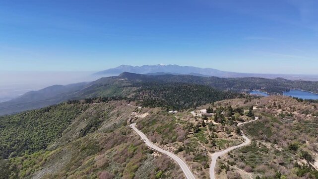 An aerial view tracing the winding asphalt of the Rim of the World Highway through dense evergreen forests toward the hazy peak of Mount Baldy.