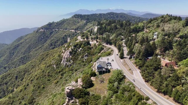 An aerial view panning left over a cliffside building on the Rim of the World Highway, revealing forested slopes with Mount Baldy and the hazy Los Angeles basin in the distance.