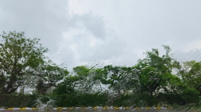 Cinematic slow motion footage of heavy raindrops hitting a car windshield during a rainstorm, with soft green tree silhouettes visible in the background. Captured in ultra high definition 4K at 240fps