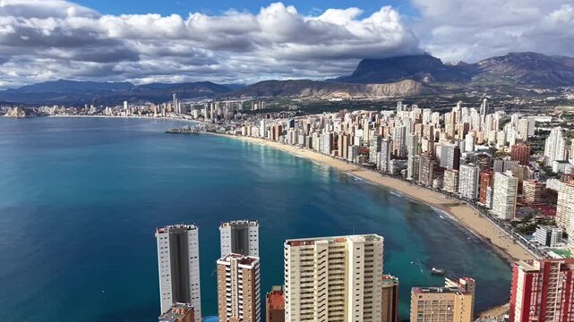 Aerial turquoise bay and skyline with highrise buildings lining sandy beach under dramatic clouds, mountain