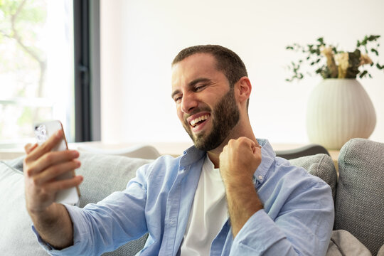 Happy Man Celebrating While Video Calling On Smartphone At Home Couch
