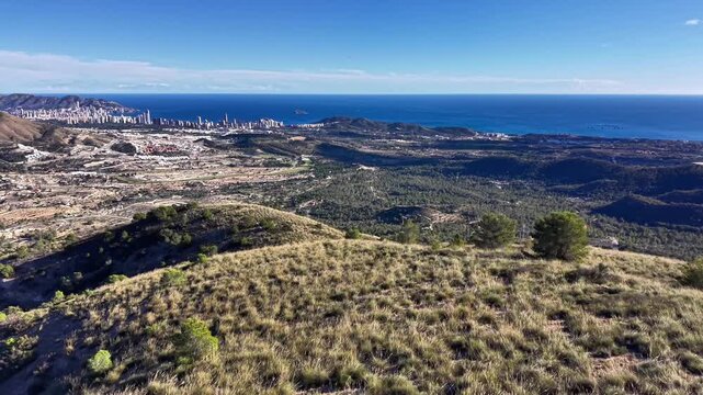 Aerial valley facing distant city skyline, golden grass slopes, scattered shrubs, textured terrain, azure sea,