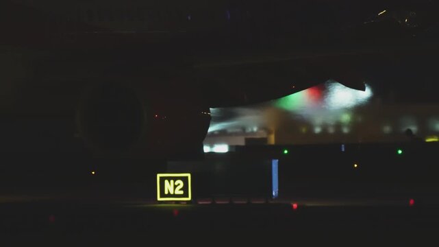 An aircraft wing with navigation and landing lights glowing cuts through the airport darkness, an illuminated taxiway sign anchoring the composition amid colorful bokeh from runway lights