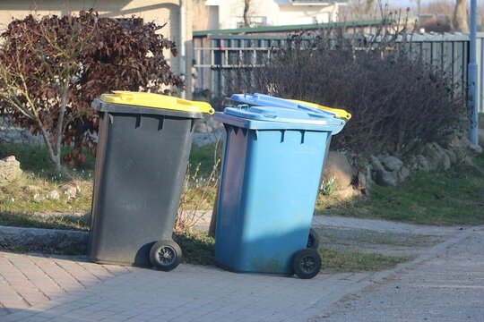 differently colored garbage cans, one blue and one dark with a yellow lid, stand on the street 
