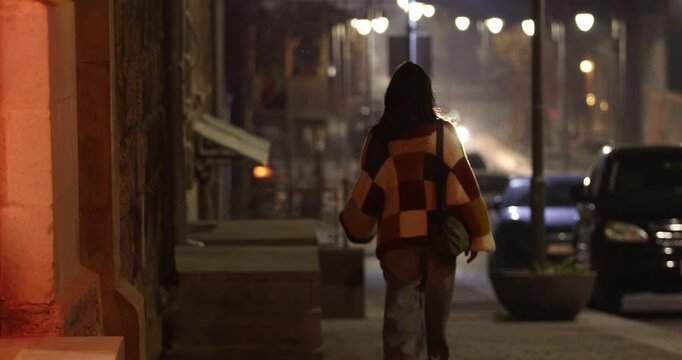 A young woman wearing a checkered sweater walks down a dimly lit, atmospheric city street at night, with bokeh streetlights and parked cars visible in the background, cinematic.