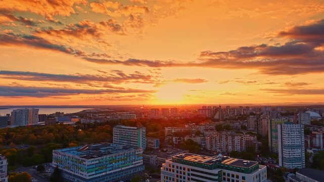 The sky displays bright colors as the sun sets over a city. Buildings are scattered throughout the landscape, and trees are visible in the foreground.