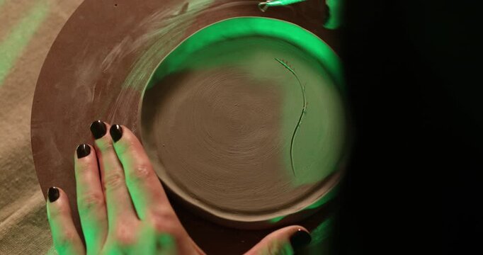 An artist meticulously carves a detailed leaf pattern into a circular clay plate using a sculpting tool, illuminated by atmospheric green lighting in a creative pottery studio