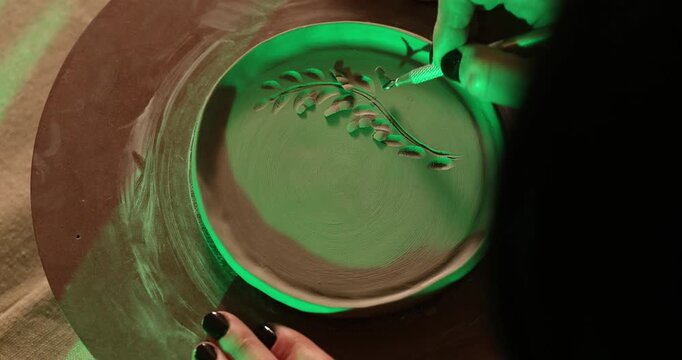 An artist meticulously carves a detailed leaf pattern into a circular clay plate using a sculpting tool, illuminated by atmospheric green lighting in a creative pottery studio