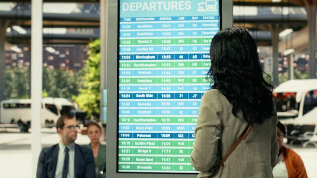 Woman passenger verifying departure time and schedule on board screen, checking timetable upon arrival at modern bus station with luggage. Traveler ready for long-distance journey.