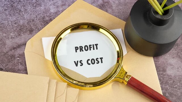Golden magnifying glass shows the phrase profit versus cost on white paper. The paper lies on a brown envelope over a textured grey surface with a dark vase