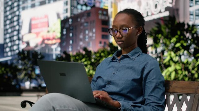 Working on laptop, sitting on bench park, digital nomad typing on keyboard and composing emails. African american woman outside writing to business partners, doing remote work.