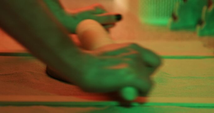 A closeup of a potter using a wooden rolling pin to flatten a disc of raw clay on a fabric-covered surface under dramatic red and green lighting.

