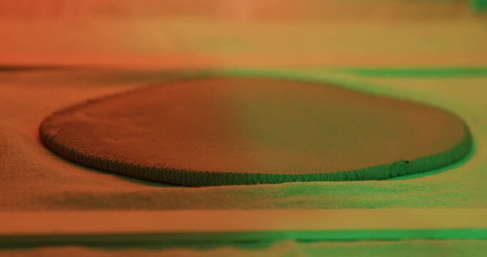 A closeup of a potter using a wooden rolling pin to flatten a disc of raw clay on a fabric-covered surface under dramatic red and green lighting.

