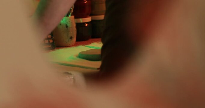A closeup of a potter using a wooden rolling pin to flatten a disc of raw clay on a fabric-covered surface under dramatic red and green lighting.


