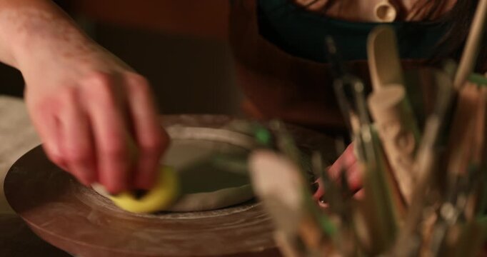 A close-up of a potter's hands using a damp yellow sponge to smooth and refine the edges of a raw clay disk on a rotating pottery wheel.

